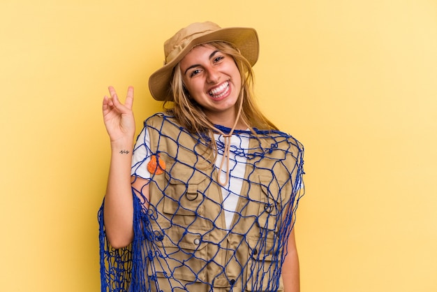 Jovem pescadora loira caucasiana segurando uma haste isolada em fundo amarelo alegre e despreocupada, mostrando um símbolo de paz com os dedos.