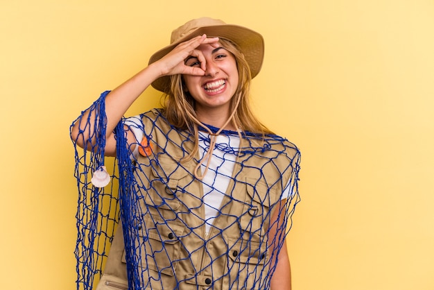 Jovem pescadora loira caucasiana segurando uma haste isolada em fundo amarelo, animada mantendo o gesto de ok no olho.