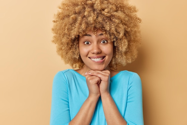 Jovem positiva com cabelo encaracolado morde os lábios mantém as mãos sob o queixo vestida com um jumper azul casual focado diretamente na câmera isolada sobre fundo bege Conceito de expressões do rosto humano