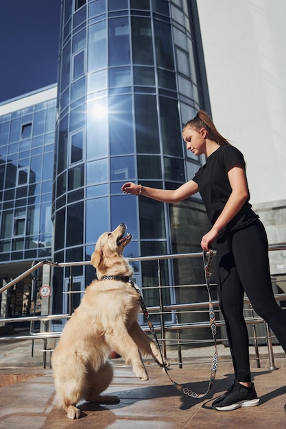 Foto jovem positiva se diverte e faz truques com seu cachorro quando caminha ao ar livre perto do prédio comercial