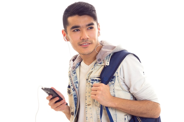 Jovem positivo com cabelo preto em camiseta branca e jaqueta jeans com mochila azul usando smartphone e fones de ouvido em fundo branco em estúdio