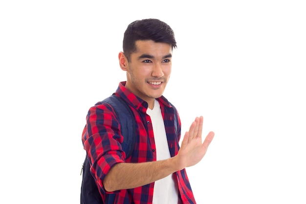 Jovem positivo com cabelo preto em camiseta e camisa xadrez vermelha com mochila acenando com a mão