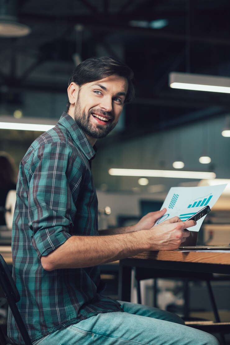 Jovem positivo com um gráfico e um smartphone sentado à mesa