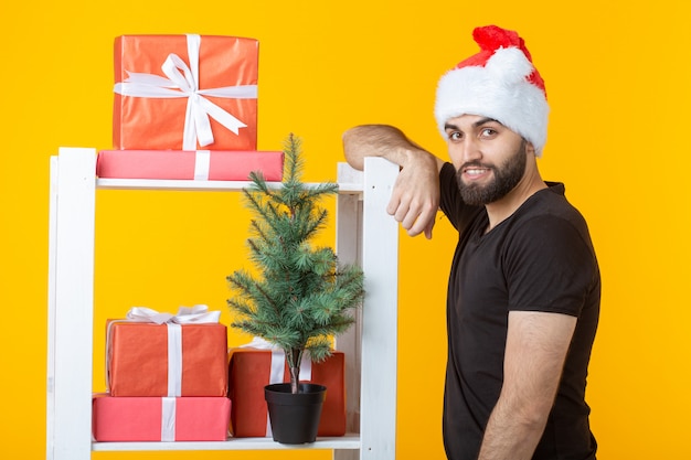 Jovem positivo com uma barba e um chapéu de Papai Noel está de pé perto da cabine com presentes e árvore de Natal em fundo amarelo. Conceito de presente de parabéns e descontos no Natal e no Ano Novo.