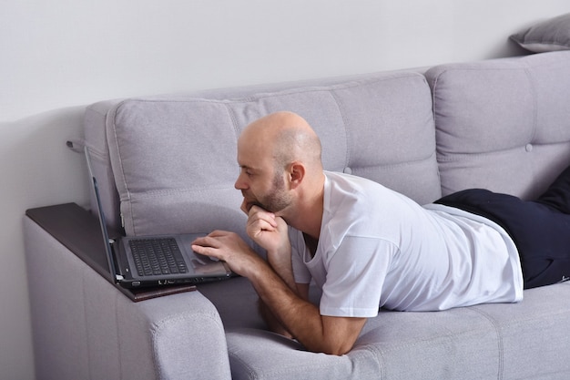 Jovem positivo em roupas casuais, sentado na sala de estar e usando um laptop