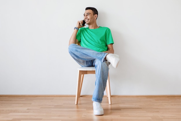 Jovem positivo falando no celular enquanto está sentado na cadeira contra a parede de luz em casa espaço de cópia grátis