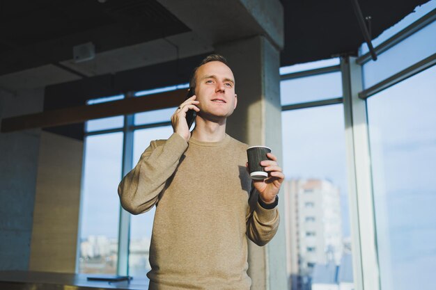 Jovem positivo sorrindo e falando no celular e bebendo café enquanto descansa em pé no escritório Pausa para o almoço durante o trabalho