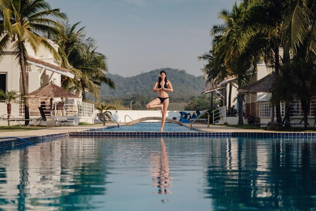 Jovem praticando ioga de pose na piscina e na praia natural ao pôr do sol ou nascer do sol Ioga e conceito saudável ioga em pose de árvore