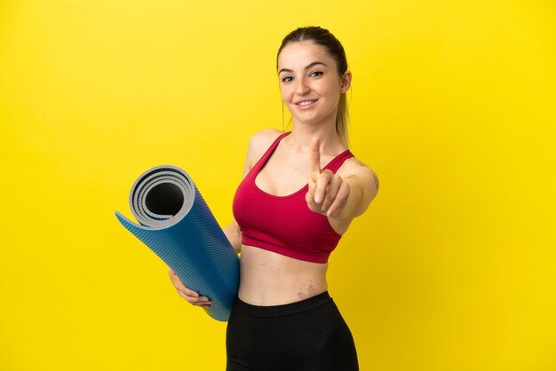 Jovem praticante de esportes indo para aulas de ioga, segurando um tapete, mostrando e levantando um dedo