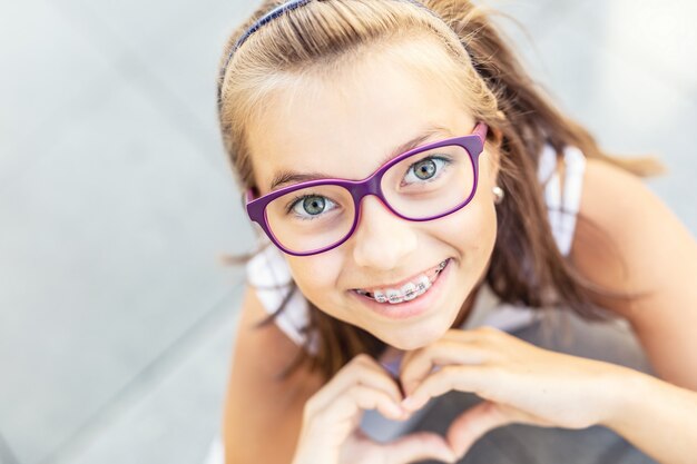 Foto jovem pré-adolescente de óculos usando aparelho sorri para a câmera, mostrando a forma do coração com as mãos.