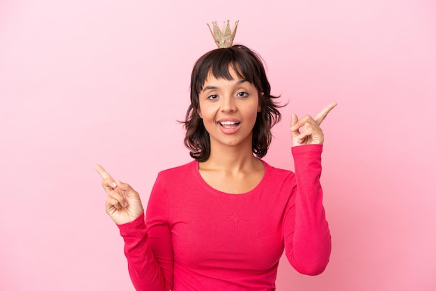 Jovem princesa mestiça com coroa isolada em fundo rosa apontando o dedo para as laterais e feliz