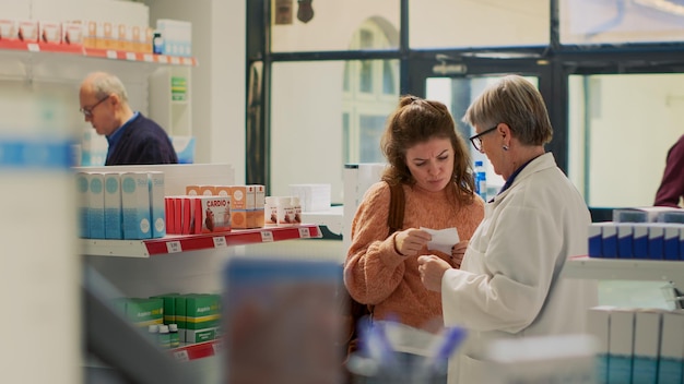 Jovem procurando suplementos em papel de prescrição, conversando com farmacêutico sobre tratamento de doenças na farmácia. Cliente verificando caixas de comprimidos e bulas.