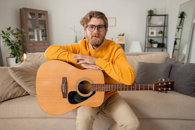 Jovem professor de música de sucesso em trajes casuais olhando para você enquanto está sentado no sofá e fazendo anotações no bloco de notas durante a aula online