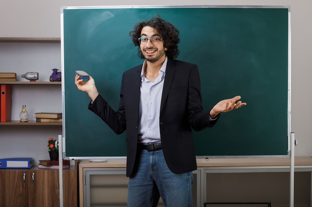 Jovem professor do sexo masculino com as mãos abertas a sorrir, de pé na frente do quadro a segurar uma esponja na sala de aula