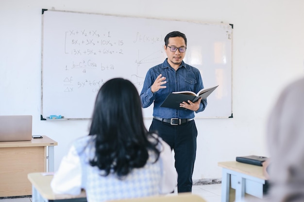 Jovem professor explicando algo na frente da sala de aula enquanto segura um livro