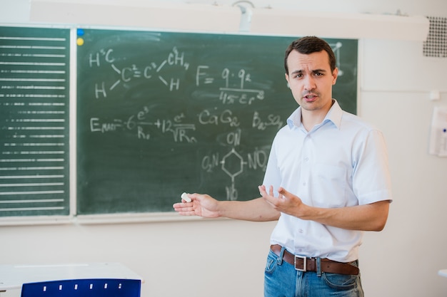 Jovem professor perto de lousa na sala de aula de escola falando com classe