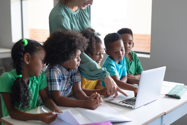 Jovem professora caucasiana apontando para o laptop enquanto ensina estudantes afro-americanos
