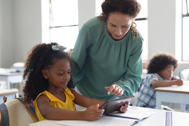 Foto jovem professora caucasiana mostrando tablet digital a uma menina afro-americana na secretária