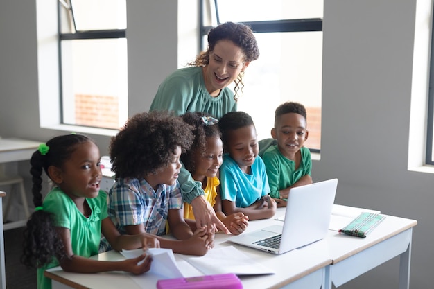 Jovem professora caucasiana olhando para estudantes primários afro-americanos enquanto ensina laptop