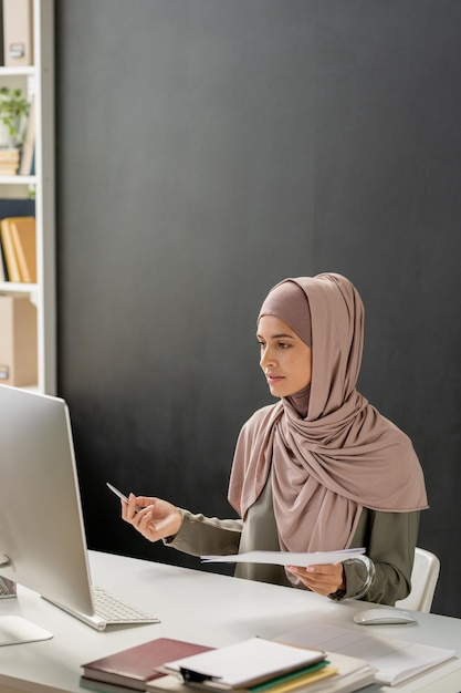 Jovem professora de hijab sentada em frente ao computador explicando o assunto da aula para o público online