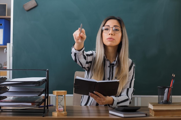 jovem professora de óculos sentado na mesa da escola na frente do quadro-negro na sala de aula verificando a lição de casa dos alunos apontando com caneta para algo ou alguém