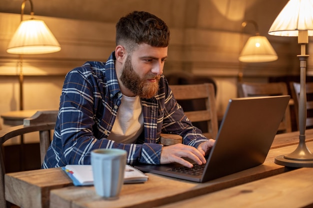 Jovem profissional navegando na Internet em seu laptop com uma xícara de café na mesa da cafeteria ou do escritório em casa, trabalhando a partir do conceito de café