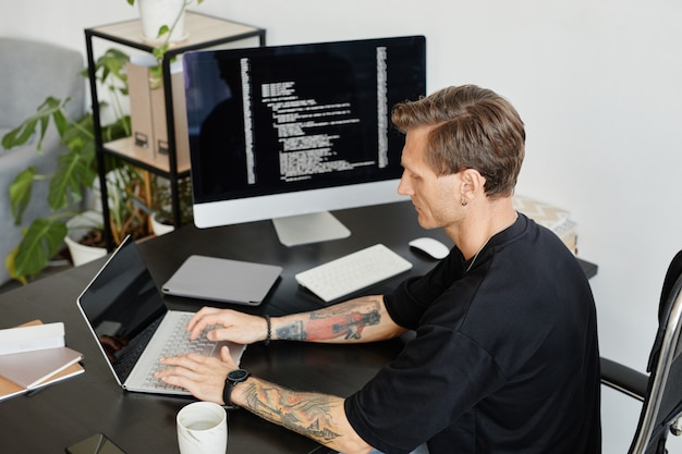 Jovem programador sentado à mesa digitando no laptop, ele desenvolvendo software no escritório