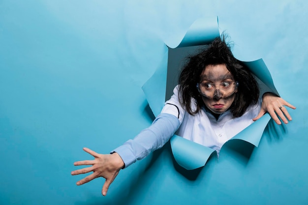 Jovem químico adulto com olhar maluco, cabelo bagunçado e cara suja agindo pateta após a explosão do experimento. Cientista humorístico tolo com expressão cômica sobre fundo azul. Tiro de estúdio.