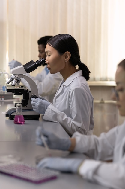 Jovem químico asiático trabalhando com microscópio na mesa enquanto trabalhava no laboratório