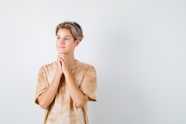 Jovem rapaz adolescente apertando as mãos sob o queixo, olhando para longe em t-shirt e parecendo um sonho, vista frontal.