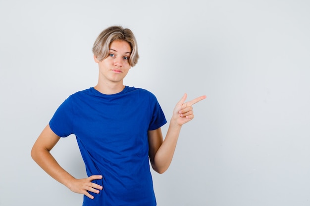 Jovem rapaz adolescente apontando para o canto superior direito em uma camiseta azul e olhando hesitante, vista frontal.