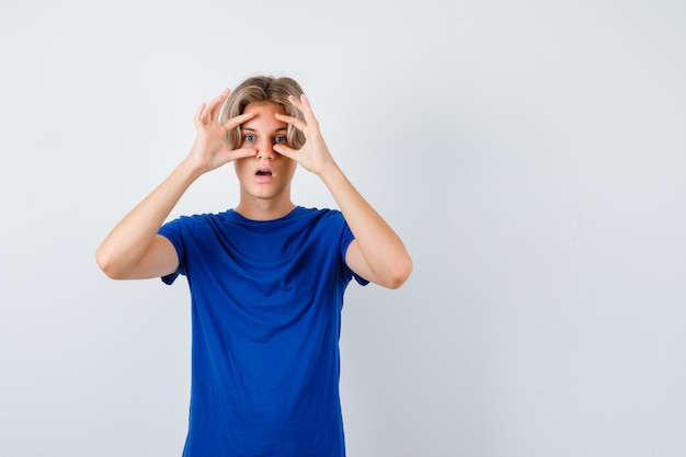 Jovem rapaz adolescente espiando por entre os dedos em uma camiseta azul e parecendo maravilhado. vista frontal.