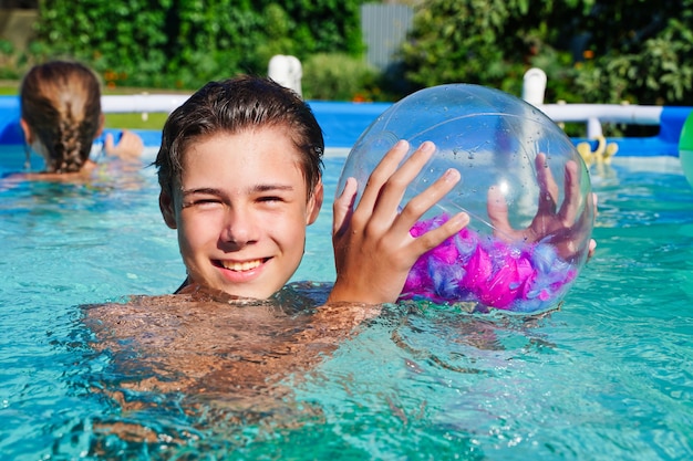 Jovem rapaz bonito sorrindo em pé na piscina com uma bola, horário de verão, dia ensolarado e água límpida