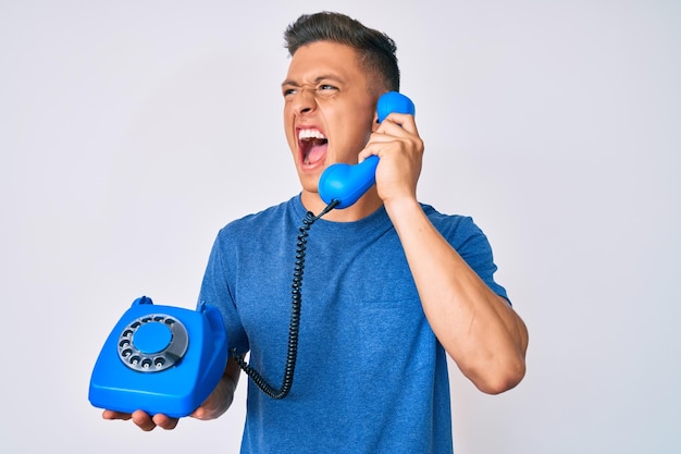 Jovem rapaz hispânico segurando telefone vintage com raiva e grito louco grito frustrado e furioso com raiva raiva e conceito agressivo