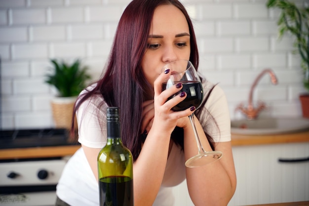 Jovem relaxada bebe vinho tinto em pé na mesa da cozinha Morena adulta descansando com álcool na cozinha