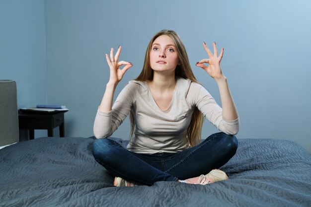 Jovem relaxada sentada em casa na cama em posição de lótus, meditando garota praticando ioga