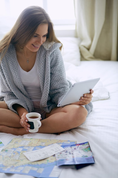 Jovem relaxada sentada na cama com uma xícara de café e tablet digital