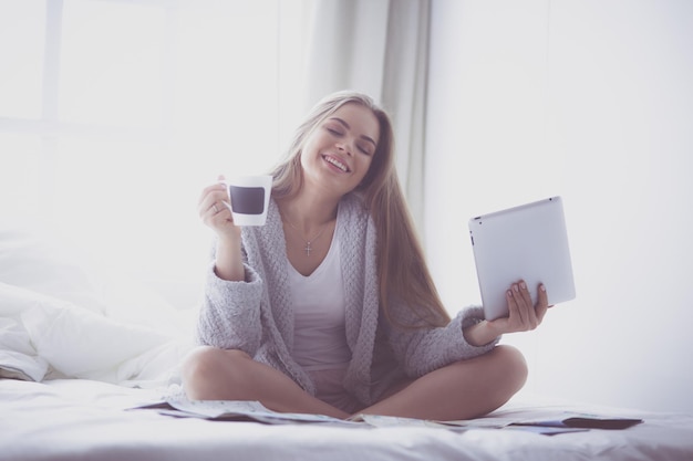 Jovem relaxada sentada na cama com uma xícara de café e tablet digital