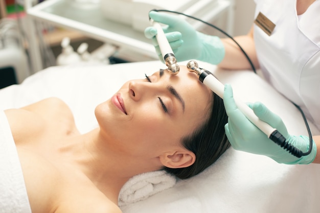 Jovem relaxando com os olhos fechados e sorrindo enquanto médico profissional com luvas de borracha segurando pinos durante a terapia de microcorrente