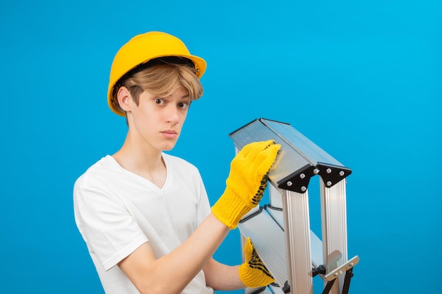 Jovem reparador adolescente em camiseta branca e luvas com capacete amarelo na cabeça segurando escada nas mãos em pé no estúdio em fundo azul Um futuro arquiteto isolado em um fundo azul