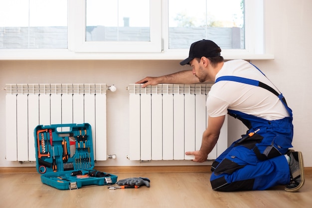 Jovem reparador profissional em uniforme especial com ferramentas está instalando radiadores e termostato em uma sala doméstica