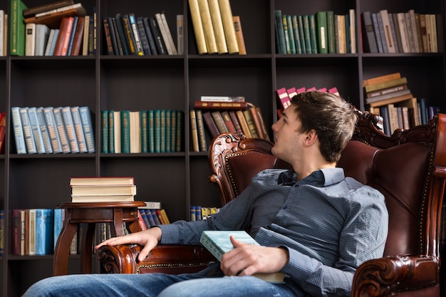 Jovem retirando livros em uma biblioteca sentado em uma poltrona confortável lendo os títulos dos livros nas prateleiras