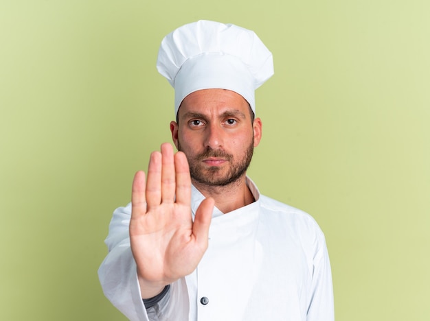 Jovem rigoroso, caucasiano, cozinheiro, com uniforme de chef e boné, olhando para a câmera, fazendo gesto de parada isolado na parede verde oliva