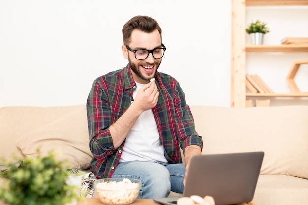 Jovem rindo com pipoca e chá enquanto assiste um filme no laptop