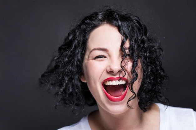 Jovem rindo contagiante com a boca aberta Linda morena encaracolada brilhante em uma regata branca com batom vermelho Fundo preto Closeup