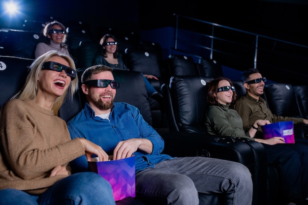 Jovem rindo, homem e mulher e seus amigos em óculos 3D, comendo pipoca e assistindo filme no cinema