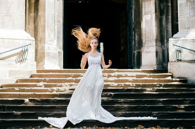 Jovem romântica e elegante com cabelo esvoaçante em um vestido longo branco posando nas escadas do antigo palácio