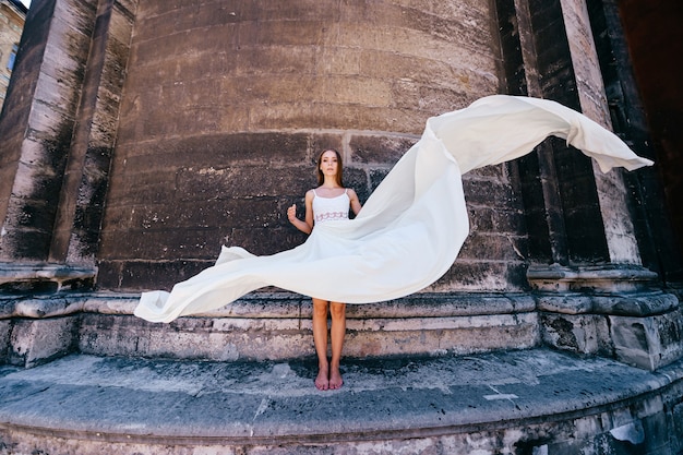 Jovem romântica e elegante em um vestido longo branco esvoaçante posando sobre uma parede de pedra antiga