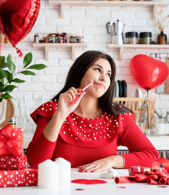 jovem romântica em um vestido vermelho escrevendo uma carta de amor sentada na cozinha decorada