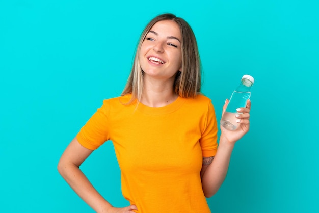 Jovem romena com uma garrafa de água isolada em fundo azul posando com braços no quadril e sorrindo